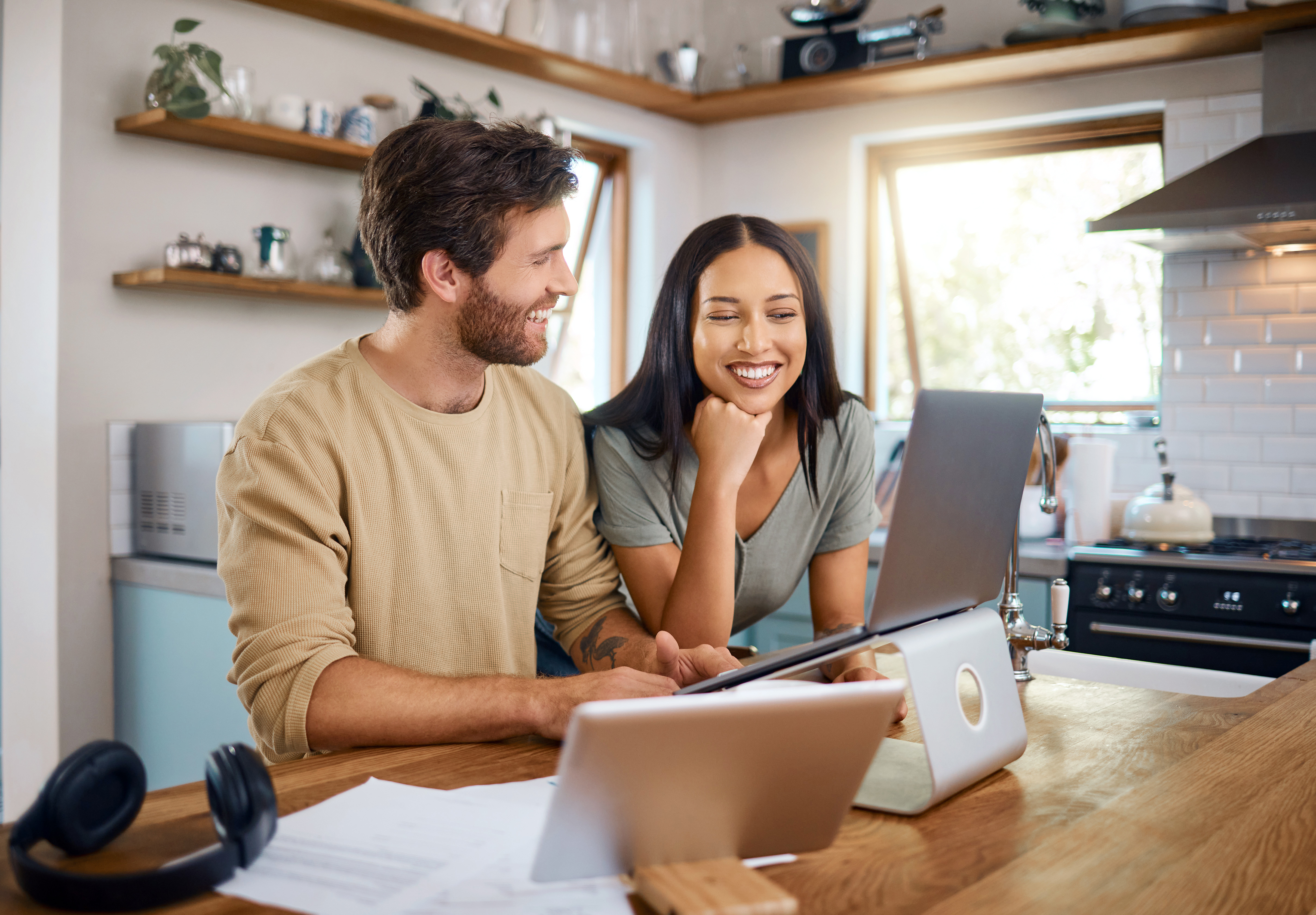 Happy young caucasian man working on laptop while his wife stands next to him looking at the screen. Man doing freelance work and getting distracted by beautiful wife | Source: Getty Images